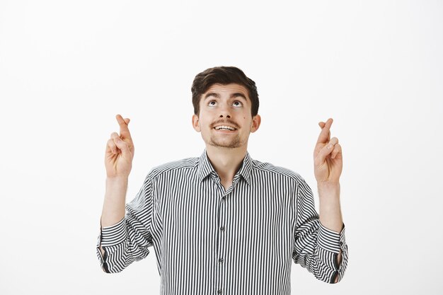 Sperando gioioso attraente ragazzo barbuto in camicia a righe, alzando lo sguardo mentre si incrociano le dita e facendo o pregando con successo, sentendosi fiducioso mentre prega per il miglior risultato sul muro grigio