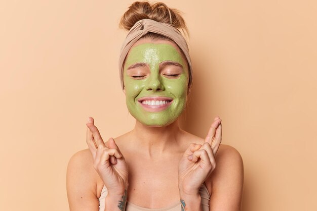 Speranzosa donna allegra incrocia le dita per buona fortuna tiene gli occhi chiusi sorrisi applica piacevolmente maschera facciale spa verde per ridurre le linee sottili indossa fascia sta in piedi spalle nude isolate su beige