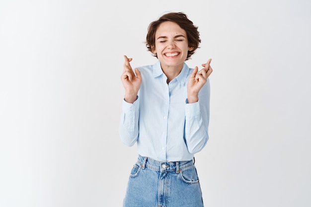 Speranzosa donna sorridente chiude gli occhi e incrocia le dita per buona fortuna, esprimendo desideri o sognando ottimisti, in piedi su un muro bianco