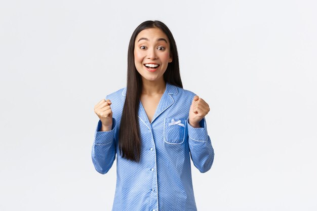 Speranzosa ragazza asiatica eccitata in pigiama blu stringe i pugni e guarda gioire della macchina fotografica, sorridendo in attesa di grandi notizie, sentendosi entusiasta e ottimista come in piedi su sfondo bianco