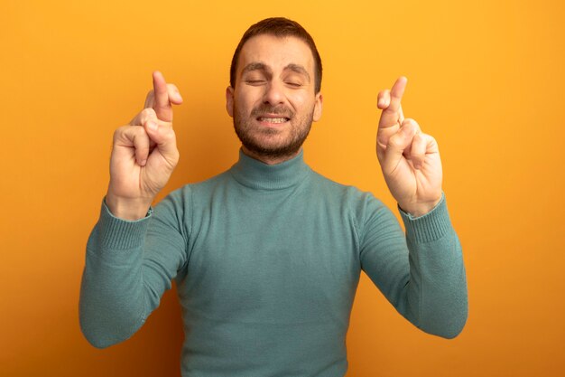 Speranzoso giovane uomo caucasico che fa gesto di buona fortuna con gli occhi chiusi isolati sulla parete arancione