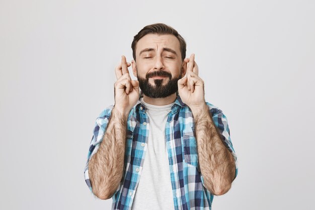 Speranzoso uomo adulto barbuto che esprime desiderio, incrociare le dita e chiudere gli occhi