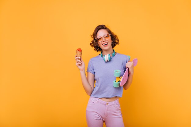 Spettacolare donna bianca con i capelli corti in posa con il gelato. Raffinata ragazza europea con skateboard isolato.