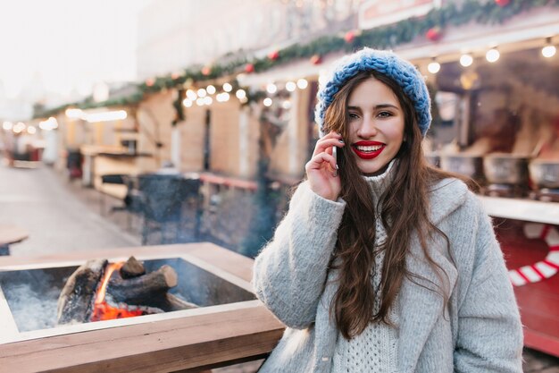 Spettacolare donna castana in cappotto grigio di lana in posa alla fiera di Natale con un sorriso. Ragazza romantica con acconciatura lunga indossa cappello blu in piedi sulla strada decorata per le vacanze invernali.