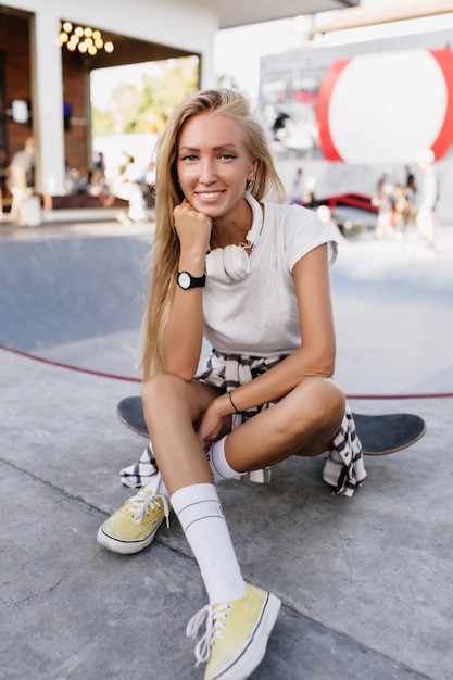 Spettacolare donna in orologio da polso nero in posa sognante sulla strada. Bella donna pattinatore seduto su skateboard con un sorriso sincero.