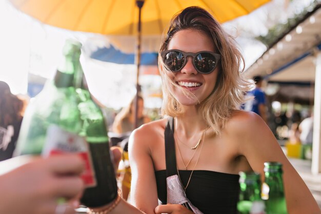 Spettacolare giovane donna che celebra qualcosa nella caffetteria estiva. Foto all'aperto di bella ragazza bionda che beve birra con gli amici in una giornata calda.