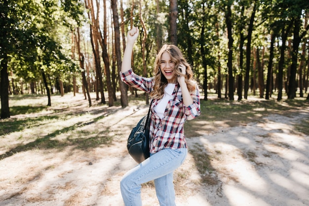 Spettacolare giovane donna in jeans che balla nella foresta estiva. Ragazza spensierata con lo zaino che scherza durante il servizio fotografico all'aperto.