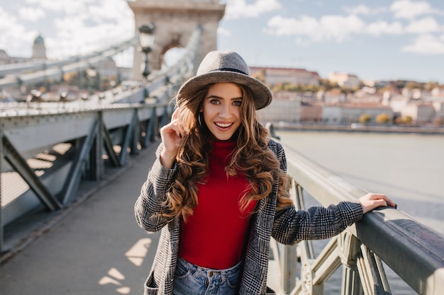 Spettacolare modello femminile bianco in cappello in posa sullo sfondo del fiume e sorridente