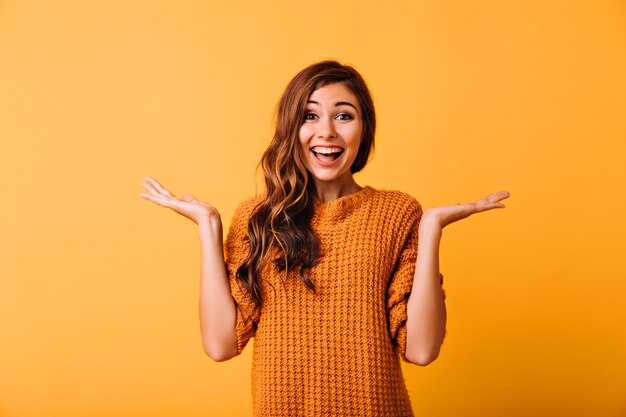 Spettacolare ragazza divertente che esprime emozioni positive su giallo. Ritratto in studio di una magnifica donna dai capelli lunghi in maglione elegante.