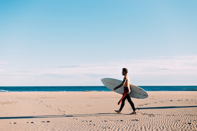 Spiaggia aperta e vuota con surfista solitario in muta a piedi verso la riva del mare o dell'oceano con la tavola da surf
