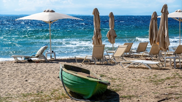 Spiaggia sulla costa del Mar Egeo con ombrelloni e lettini, barca spiaggiata in metallo di colore verde a Nikiti, Grecia