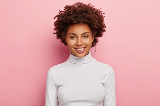 Splendida bella donna ha un sorriso piacevole sul viso, pelle sana, taglio di capelli afro, indossa un maglione dolcevita bianco casual