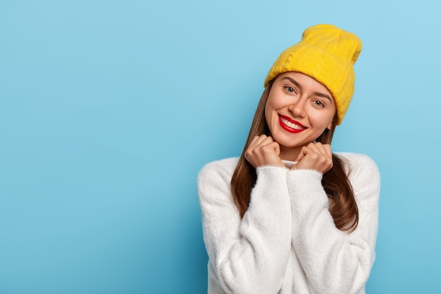 Splendida bella giovane donna inclina la testa, ha un aspetto tenero, sorride felice, indossa rossetto rosso, trucco minimo, vestito con cappello giallo e maglione bianco comodo, isolato su sfondo blu