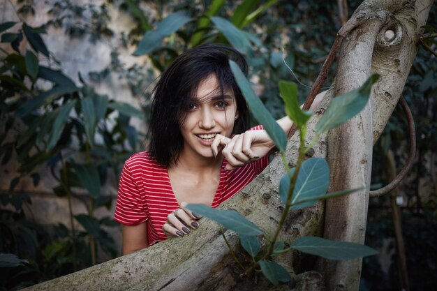 Splendida bella ragazza che fissa la telecamera con un sorriso giocoso, toccando misteriosamente le labbra mentre si cammina in giardino o parco, circondato da piante esotiche. Natura, persone, stile di vita e concetto di tempo libero