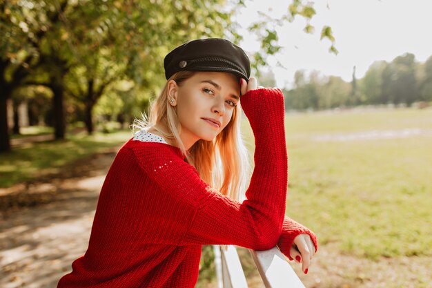 Splendida bionda che gode del tempo soleggiato all'aperto. Bella ragazza in pullover rosso alla moda che sembra buono nel parco d'autunno.