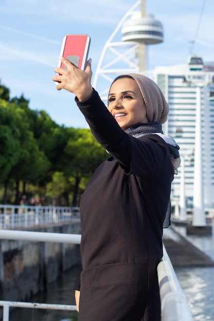 Splendida donna araba sorridente che prende selfie. Donna con la testa coperta e trucco immaginandosi con il telefono cellulare. Concetto di social media internazionale, bello