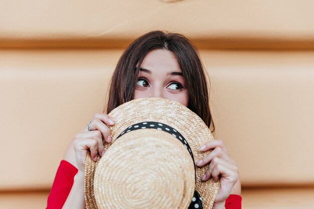 Splendida donna caucasica con cappello estivo in mano scherzare all'aperto. Signora castana con l'espressione del viso felice in posa davanti al muro.