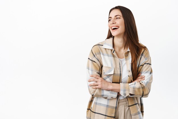 Splendida donna che ride, guardando da parte il testo promozionale dello spazio copia con un sorriso compiaciuto, in piedi in camicia a quadri sul muro bianco