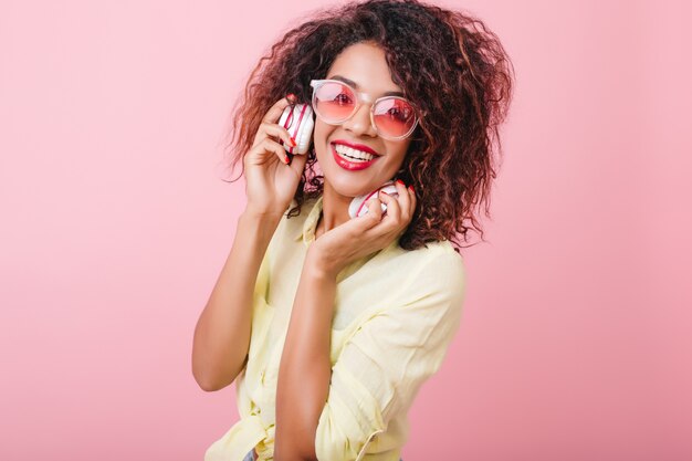 Splendida donna con la pelle lucida marrone chiaro rilassata con la musica preferita. ritratto di ragazza mulatta felice in occhiali da sole colorati alla moda ascoltando musica in cuffia.