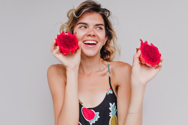 Splendida donna dai capelli corti in posa con un sorriso ispirato e mangiare pitahaya. Tiro al coperto di attraente signora abbronzata che tiene i frutti del drago.