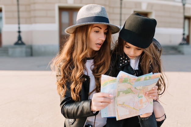 Splendida donna dai capelli lunghi guardando la mappa con l'espressione del viso confuso. Ritratto di bambina che indossa un cappello nero, pianificazione del percorso intorno alla città sconosciuta con sua madre.