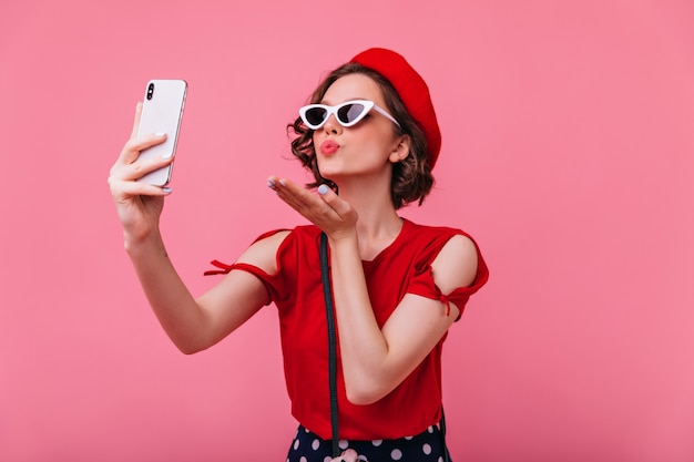 Splendida donna francese che manda un bacio d'aria mentre si scatta una foto. Ritratto dell'interno della signora riccia romantica in betet che fa selfie.