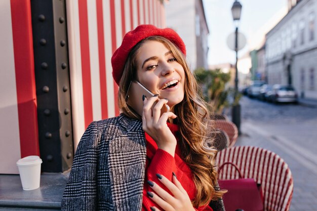 Splendida donna francese con lunghi capelli castani che parla al telefono in un caffè all'aperto. Ritratto di una ragazza riccia a riposo con un berretto rosso che chiama qualcuno sullo sfondo della città sfocata.