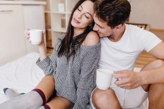 Splendida donna in maglione di lana in posa con gli occhi chiusi, seduta a letto con il fidanzato