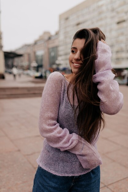 Splendida donna sorridente con i capelli lunghi in posa con piacere in città su sfocatura dello sfondo. Meravigliosa donna riccia con gli occhi scuri in posa giocosamente sulla strada.