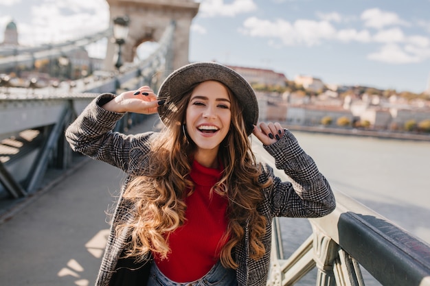 Splendida donna sorridente con lunghi capelli ricci in posa con piacere sul ponte su sfocatura dello sfondo