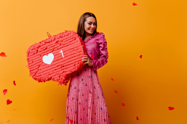 Splendida, gioiosa, allegra ragazza in posa con decorazioni rosse nelle sue mani. Ritratto di donna dai capelli castani in abito rosa contro il muro di coriandoli a forma di cuori