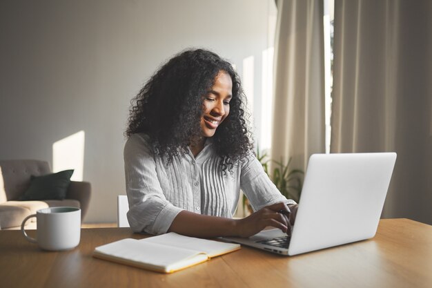 Splendida giovane blogger femmina dalla pelle scura positiva che digita su laptop generico, sorridente, ispirata durante la creazione di nuovi contenuti per il suo blog di viaggio, seduto alla scrivania con diario e tazza