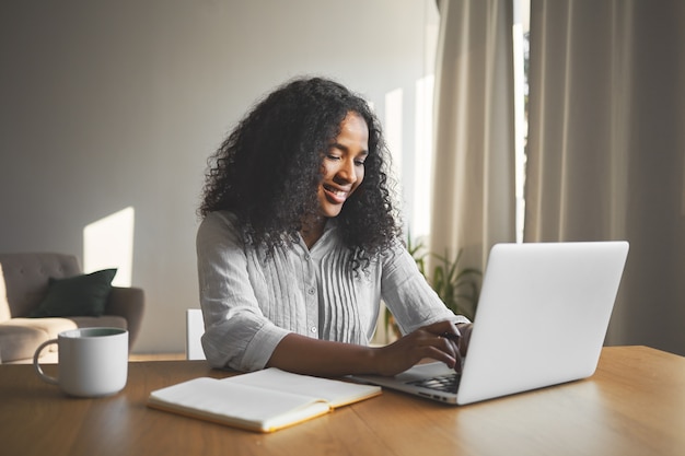 Splendida giovane blogger femmina dalla pelle scura positiva che digita su laptop generico, sorridente, ispirata durante la creazione di nuovi contenuti per il suo blog di viaggio, seduto alla scrivania con diario e tazza