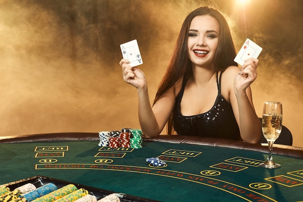 Splendida giovane donna emotiva in un vestito nero seduta al tavolo da poker con un bicchiere di champagne. Lascia che il fumo. Poker. Casinò