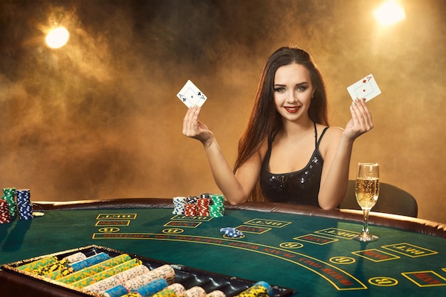 Splendida giovane donna emotiva in un vestito nero seduta al tavolo da poker con un bicchiere di champagne. Lascia che il fumo. Poker. Casinò