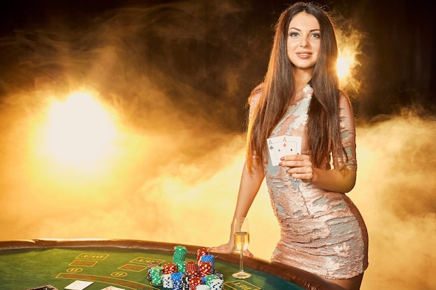 Splendida giovane donna in abito da sera con due carte in mano in piedi vicino al tavolo da poker con un bicchiere di champagne