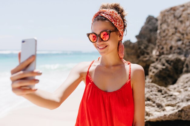 Splendida giovane donna in abito rosso utilizzando il telefono per selfie in spiaggia selvaggia. Adorabile ragazza bianca in occhiali da sole sparkle che si prende una foto mentre riposa all'oceano