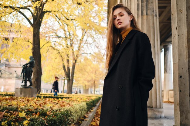 Splendida ragazza alla moda in cappotto che guarda arrogantemente a porte chiuse in piedi su un'accogliente strada autunnale