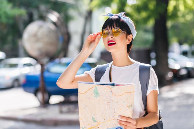 Splendida ragazza bruna in abito vintage tenendo la mappa della città e guardando in lontananza con l'espressione del viso preoccupato