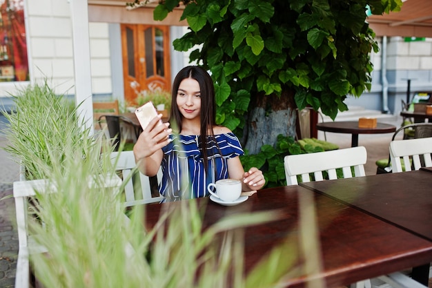 Splendida ragazza bruna seduta sul tavolo nella caffetteria con una tazza di caffè e fare selfie