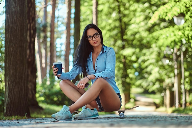 Splendida ragazza bruna sexy con gli occhiali che indossa una maglietta e pantaloncini seduto su uno skateboard e tiene una tazza di caffè da asporto e smartphone in un parco.