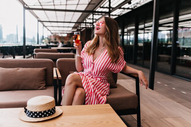 Splendida ragazza caucasica bionda che ride mentre beve vino in una giornata estiva. Foto di adorabile modello femminile in abito a righe in posa in un accogliente caffè.