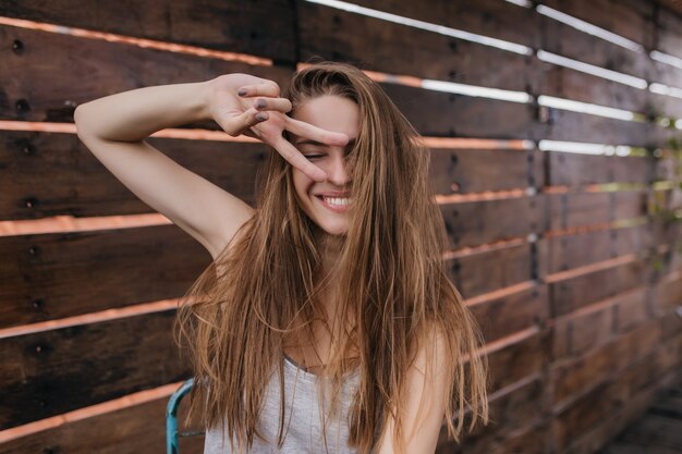 Splendida ragazza con acconciatura dritta scherzare sulla parete di legno. Foto all'aperto di giovane donna entusiasta che fa facce buffe.