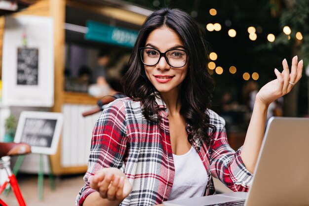 Splendida ragazza con capelli ondulati utilizzando il computer e sorridente. Ritratto all'aperto della signora caucasica che esprime interesse durante il lavoro con il computer portatile.