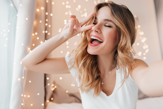 Splendida ragazza con trucco romantico in posa con gli occhi chiusi. Foto dell'interno del modello femminile attraente che fa selfie nella sua stanza.