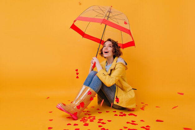 Splendida ragazza dai capelli corti con bellissimi occhi in posa sotto l'ombrellone. Foto dell'interno del modello femminile bianco romantico che si siede sul pavimento giallo con l'ombrello.
