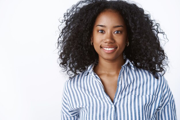 Splendida ragazza dall'aspetto amichevole sorridente felice dalla pelle scura acconciatura afro che indossa una camicetta a righe da ufficio sembra alla moda, atteggiamento ambizioso, preparati al colloquio di lavoro, in piedi muro bianco