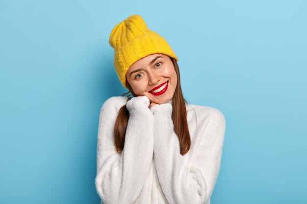 Splendida ragazza piuttosto femminile inclina la testa, indossa un morbido maglione bianco, copricapo giallo, ha le labbra rosse, esprime emozioni positive