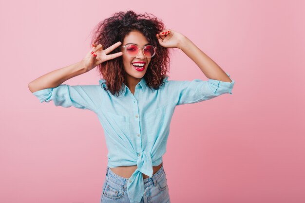 Splendida signora africana in camicia blu che esprime emozioni positive. Ritratto dell'interno di graziosa donna sorridente in occhiali da sole rosa in posa con le mani in alto.