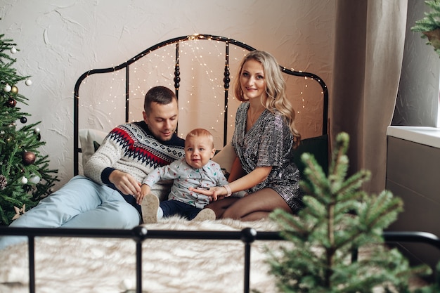 Splendida signora bionda con il suo amorevole marito e bambino carino seduto su un letto vicino all'albero di Natale